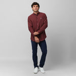 Full-length front view of a male model wearing a burgundy long-sleeve oxford shirt with a white logo, navy trousers, and white sneakers, standing against a light grey background while rolling up his sleeve