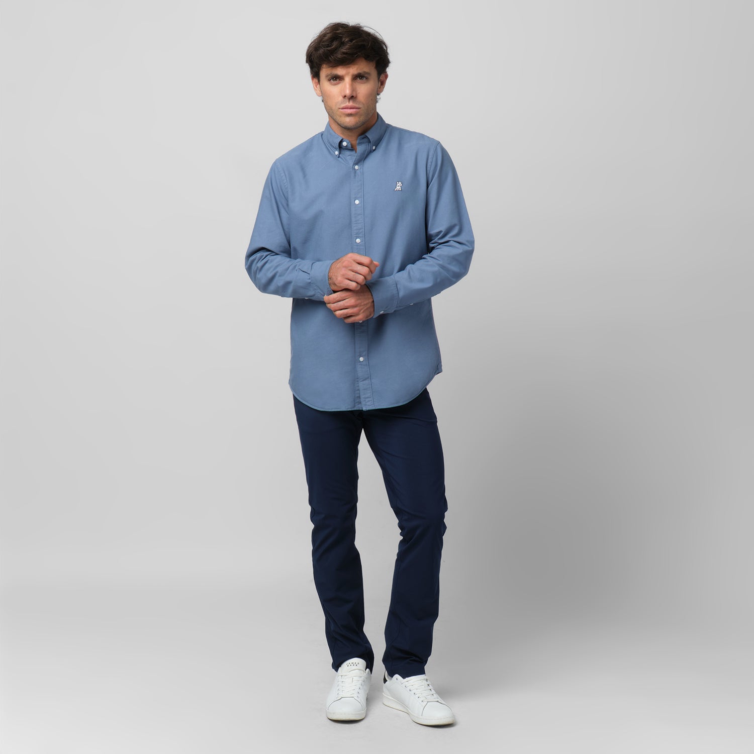 Full-length front view of a male model wearing a blue-grey long-sleeve oxford shirt with a white logo, navy trousers, and white sneakers, standing against a light grey background while adjusting his cuff