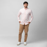 Full-length front view of a male model wearing a light pink long-sleeve oxford shirt with a pink logo, brown trousers, and white/beige sneakers, standing against a light grey background with hands in pockets