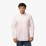 Full front view of a man wearing a pink long-sleeved button-down shirt with a pink logo and dark grey pants, standing against a white background