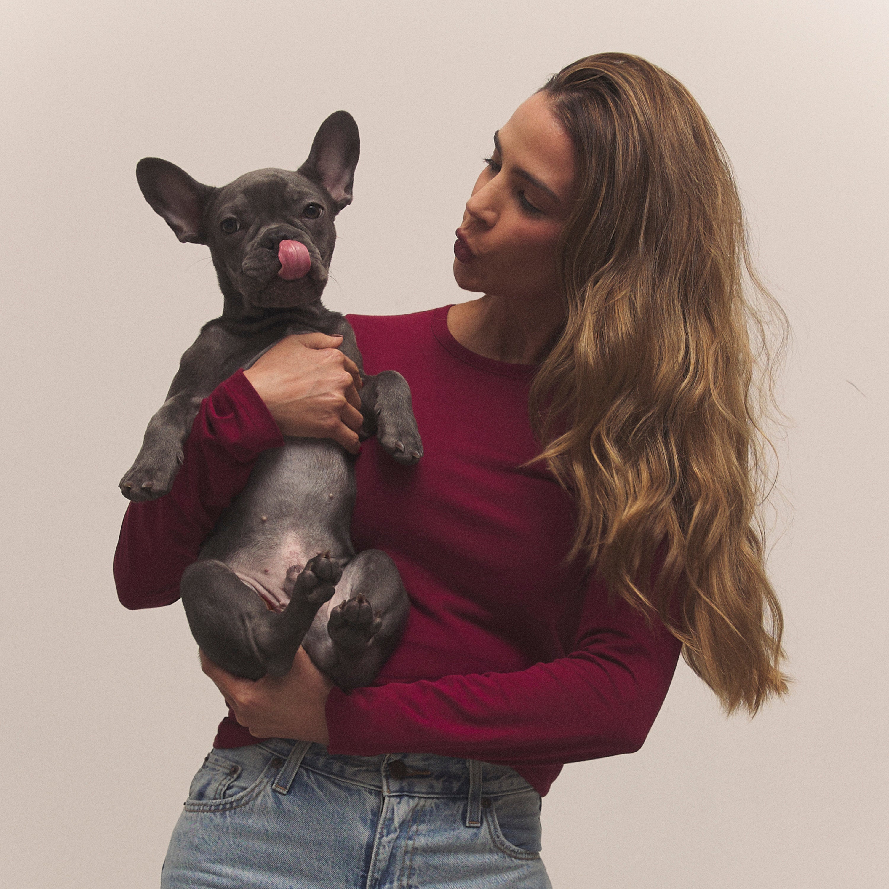 A woman in a long-sleeved burgundy shirt and blue jeans holds a small grey French Bulldog puppy that is licking its nose. She is making a kissing face towards the puppy