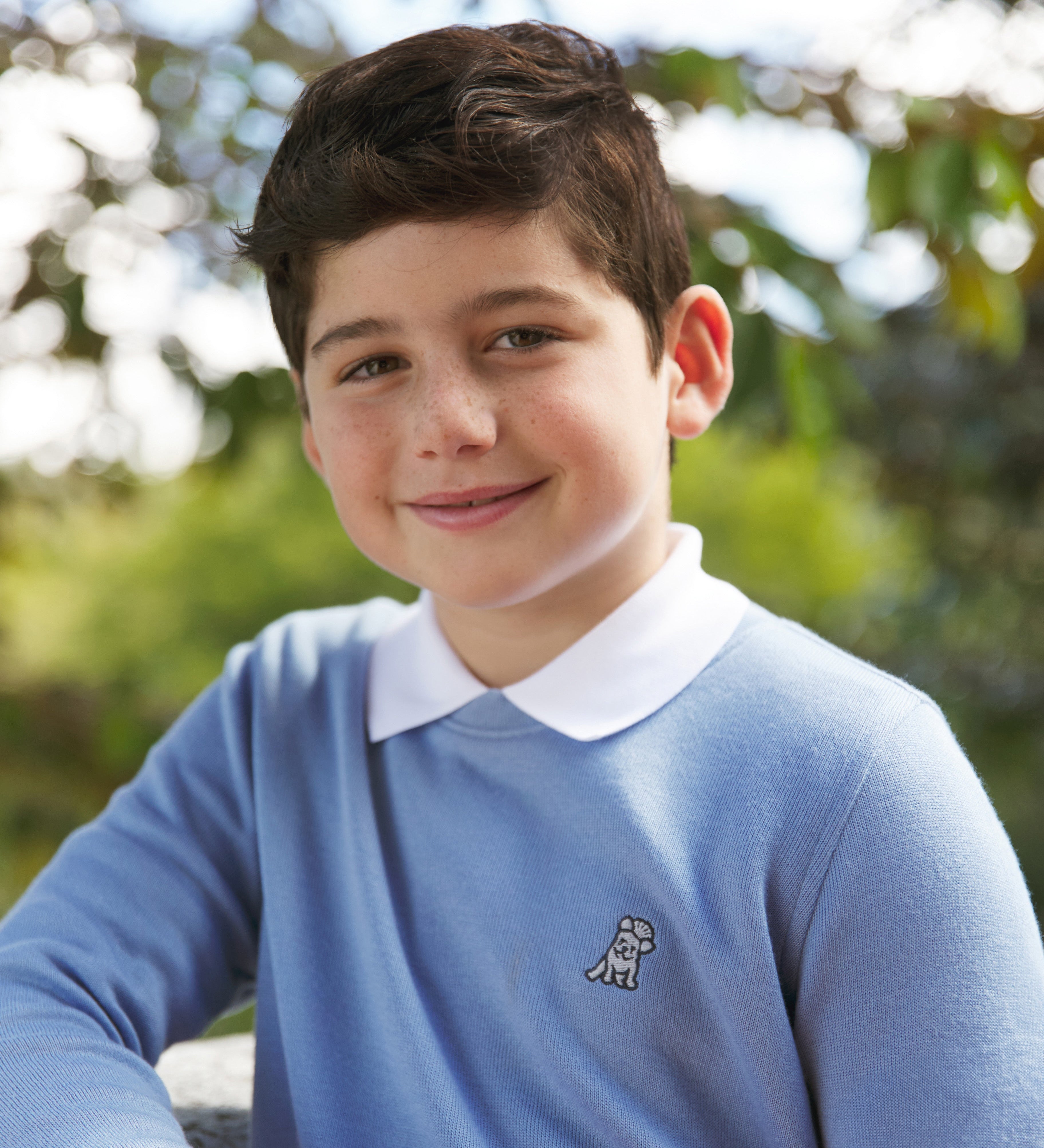Young boy wearing a light blue sweater with a white collar and logo, standing outdoors.