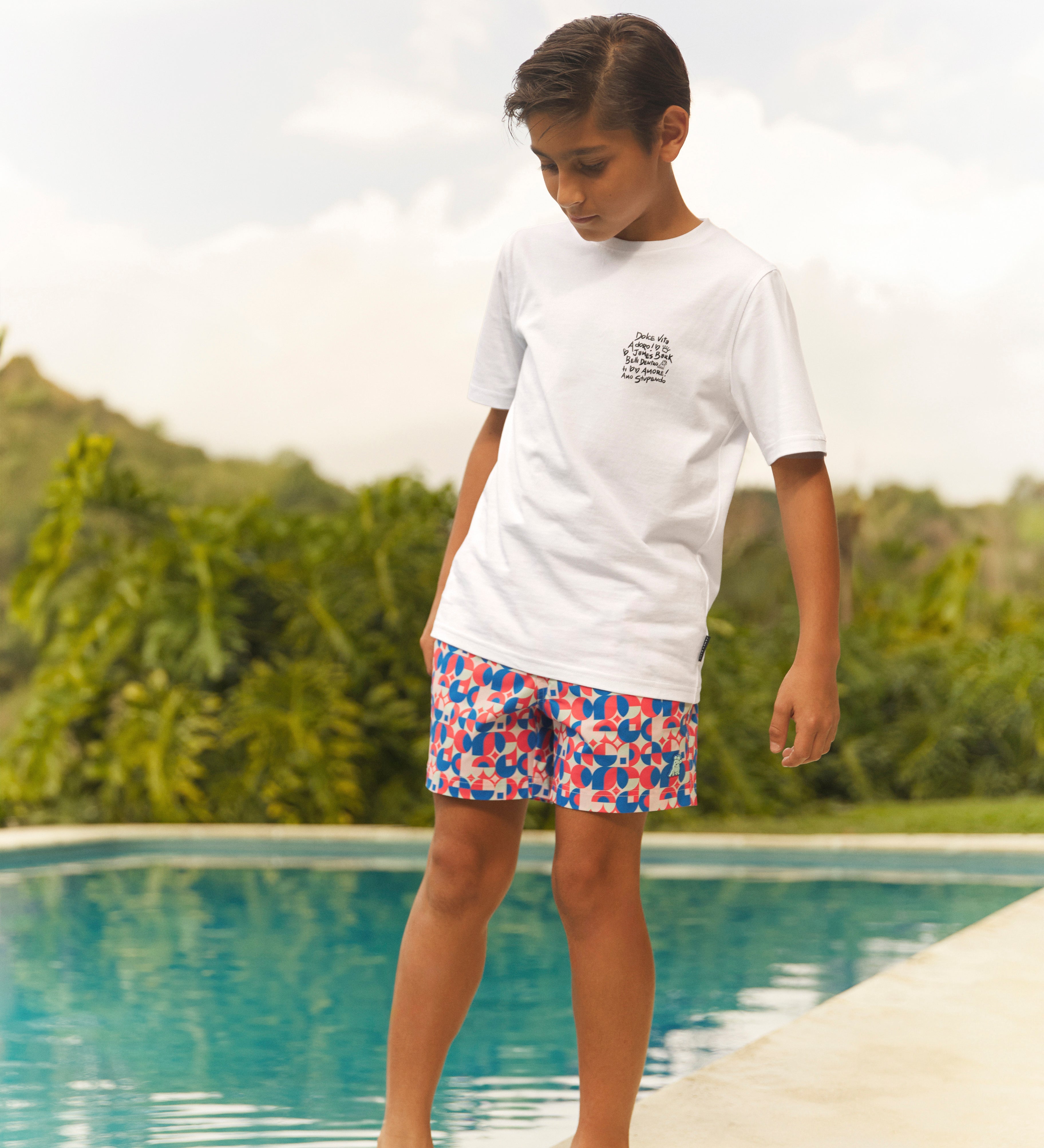 Child wearing a white t-shirt and colorful swim shorts standing by a pool with greenery in the background