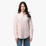 Front waist-up view of a female model wearing a light pink long-sleeve button-down oxford shirt with a small pink embroidered logo on the left chest, paired with dark wash jeans