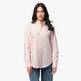 Front waist-up view of a female model wearing a light pink long-sleeve button-down oxford shirt with a small pink embroidered logo on the left chest, paired with dark wash jeans