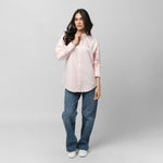 Full-length front view of a female model wearing a light pink long-sleeve oxford shirt with a pink logo, dark wash jeans, and white sneakers, standing against a light grey background and adjusting her collar