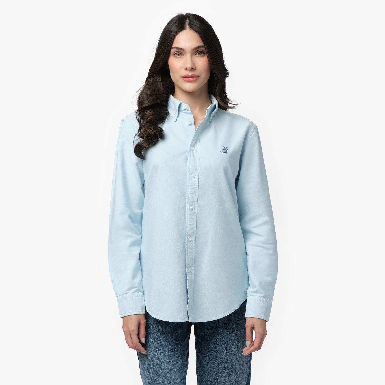 Front waist-up view of a female model wearing a light blue long-sleeve button-down oxford shirt with a small blue embroidered logo on the left chest, paired with dark wash jeans