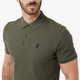 A man wearing an olive green polo shirt with a textured collar and a small dark embroidered dog logo on the left chest