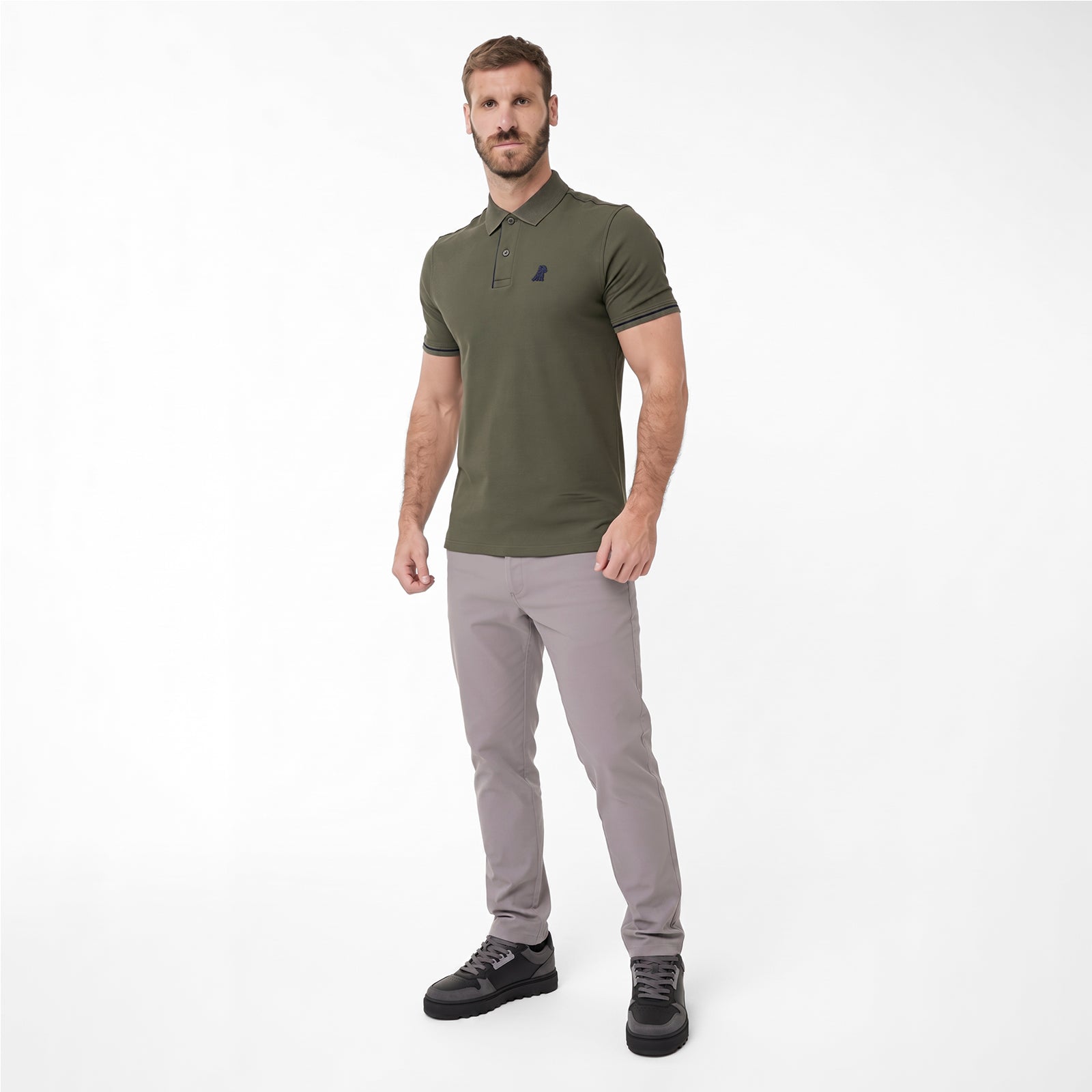 A man modeling a polo shirt in olive green with a small dark embroidered logo on the left chest, paired with light gray trousers and black sneakers