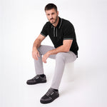 A man wearing a black polo shirt with white piping, light gray trousers, and dark gray sneakers, sitting on a white cylindrical