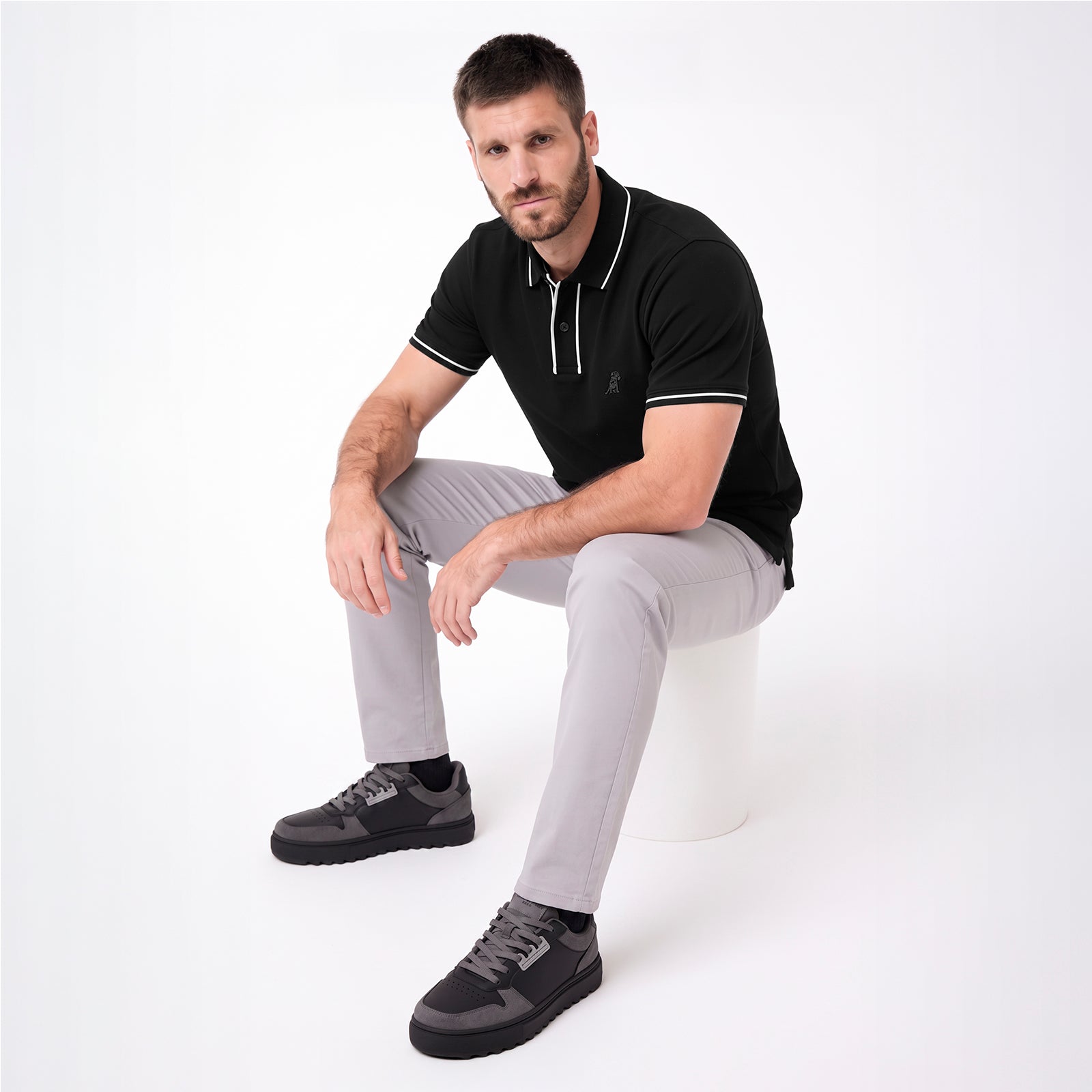 A man wearing a black polo shirt with white piping, light gray trousers, and dark gray sneakers, sitting on a white cylindrical
