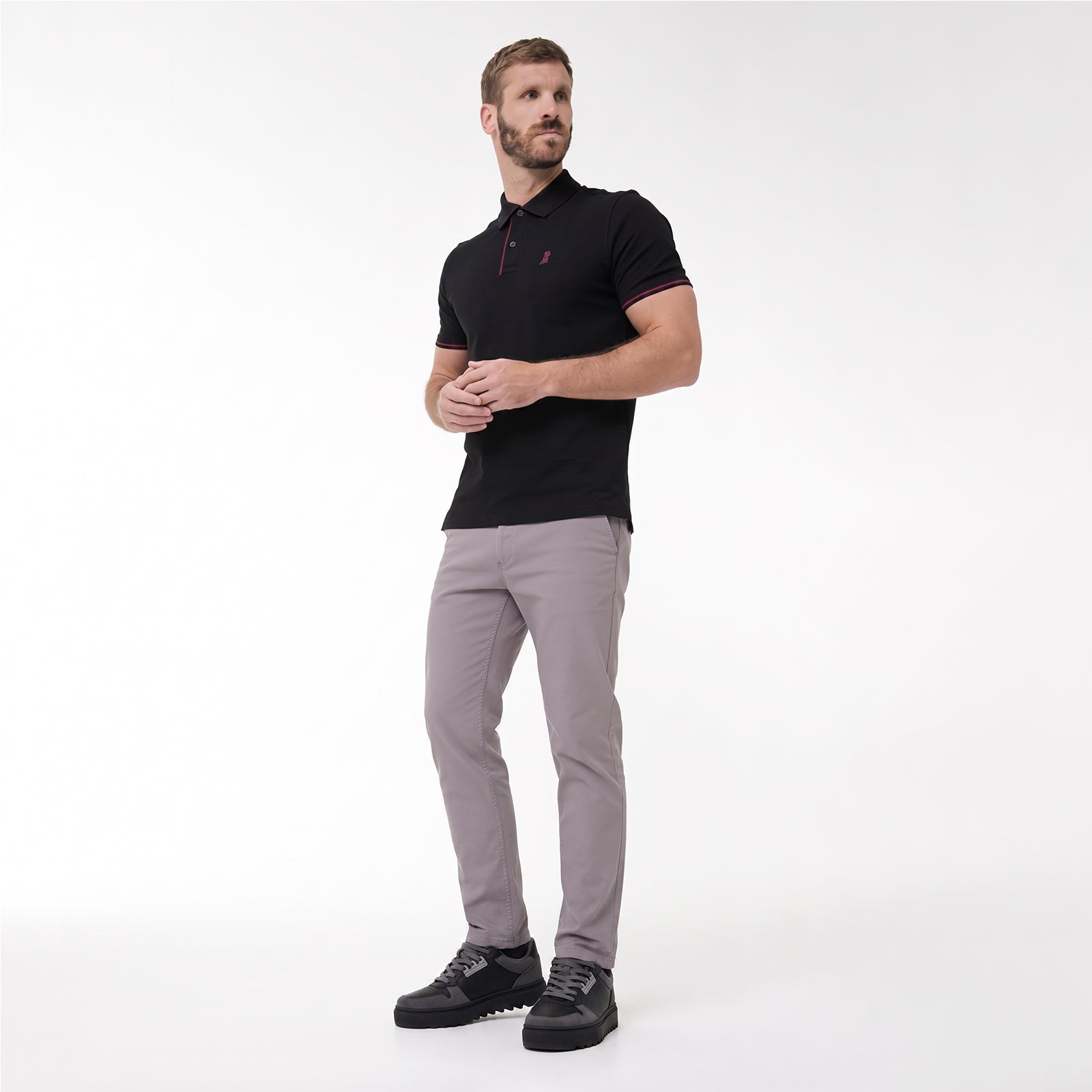 A man modeling a black polo shirt with a small red embroidered logo on the chest, paired with light gray trousers and black sneakers