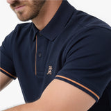 A close-up of a man wearing a navy blue polo shirt with orange contrast stitching and a small embroidered dog logo on the chest.