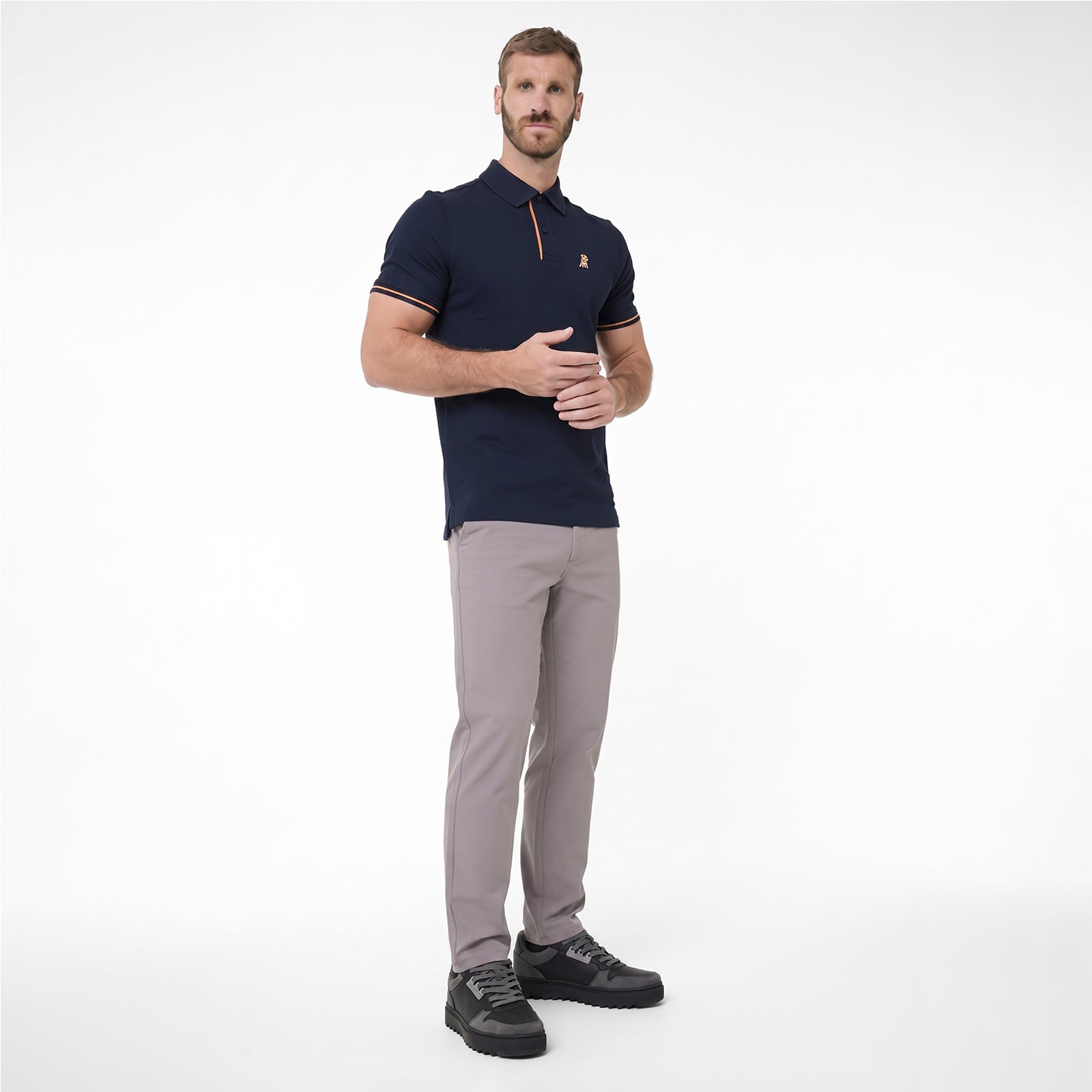 A man modeling a navy blue polo shirt with a small gold embroidered logo on the chest, paired with light gray trousers and black sneakers