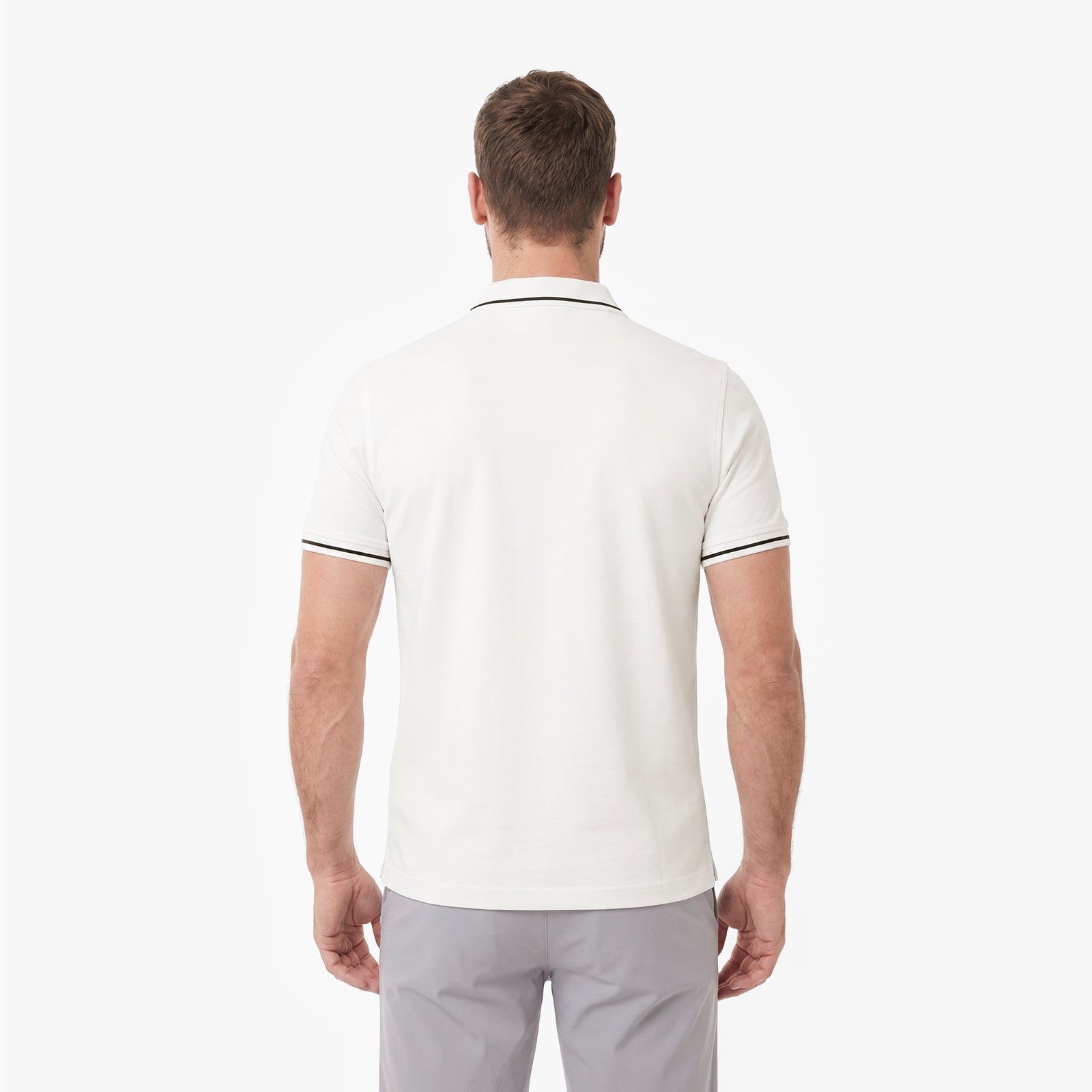 A man viewed from behind, wearing a polo shirt in off-white with a thin black stripe trim on the collar and sleeve cuffs, paired with light gray trousers