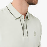 A close-up of a man wearing a light sage green polo shirt with dark contrast piping and a small embroidered dog logo on the chest
