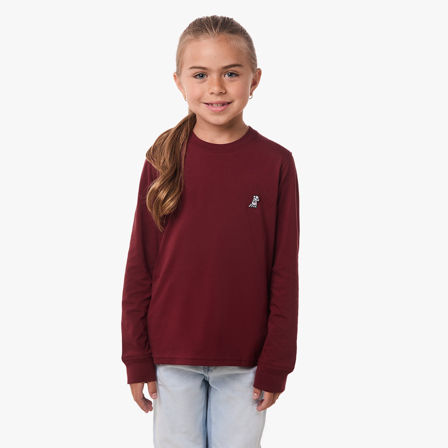 A young girl wearing a burgundy long-sleeve shirt with a small embroidered logo on the chest, standing and smiling.