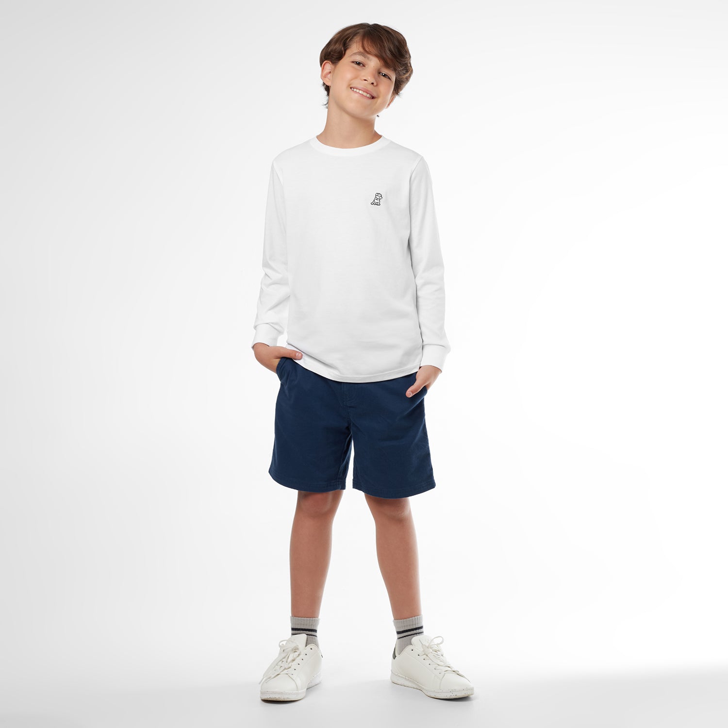 Full picture of a young boy wearing a white long-sleeved t-shirt with a small dog logo and navy shorts, standing front view