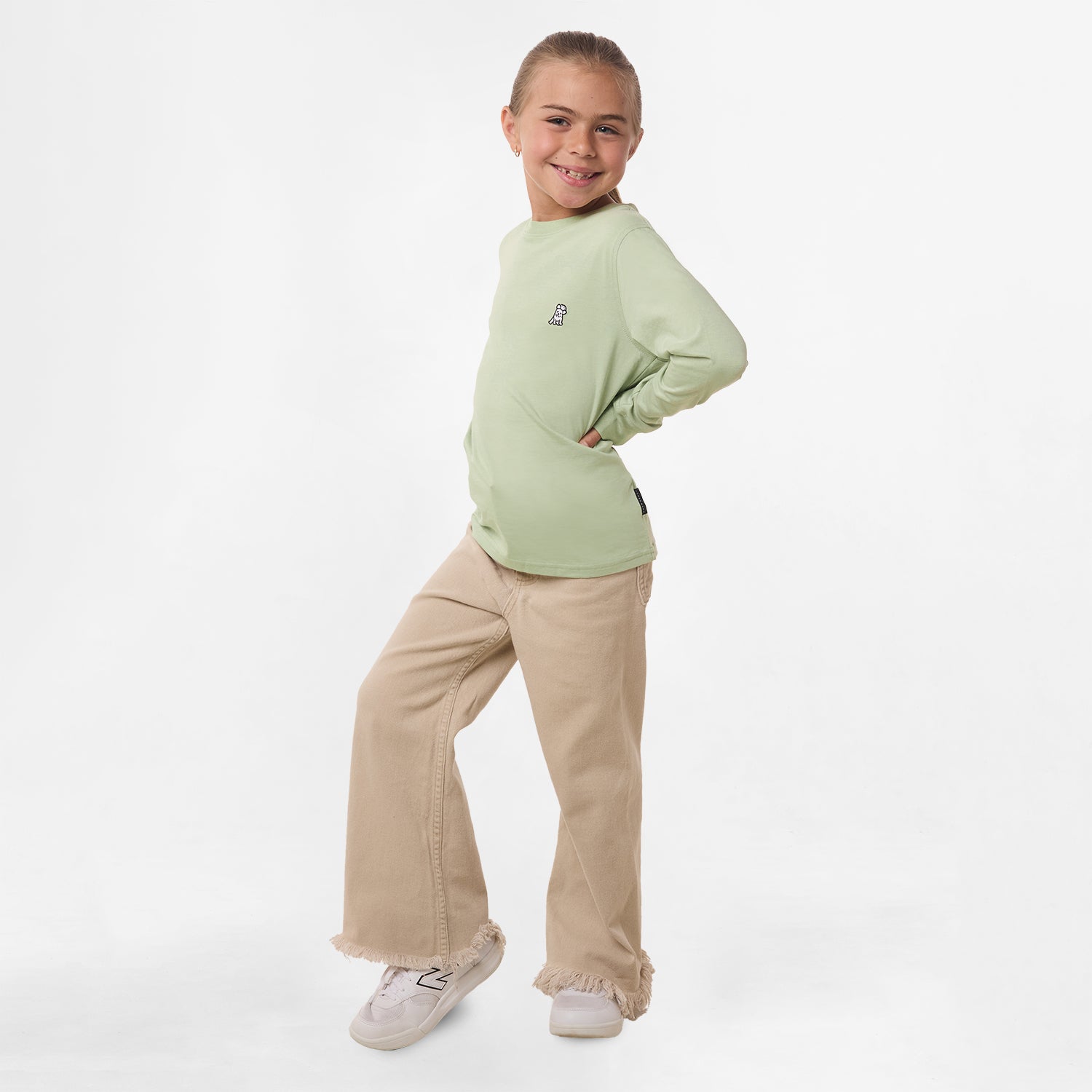 Full-body shot of the girl wearing the light green long-sleeve shirt paired with beige pants, standing with her hand on her hip and smiling.