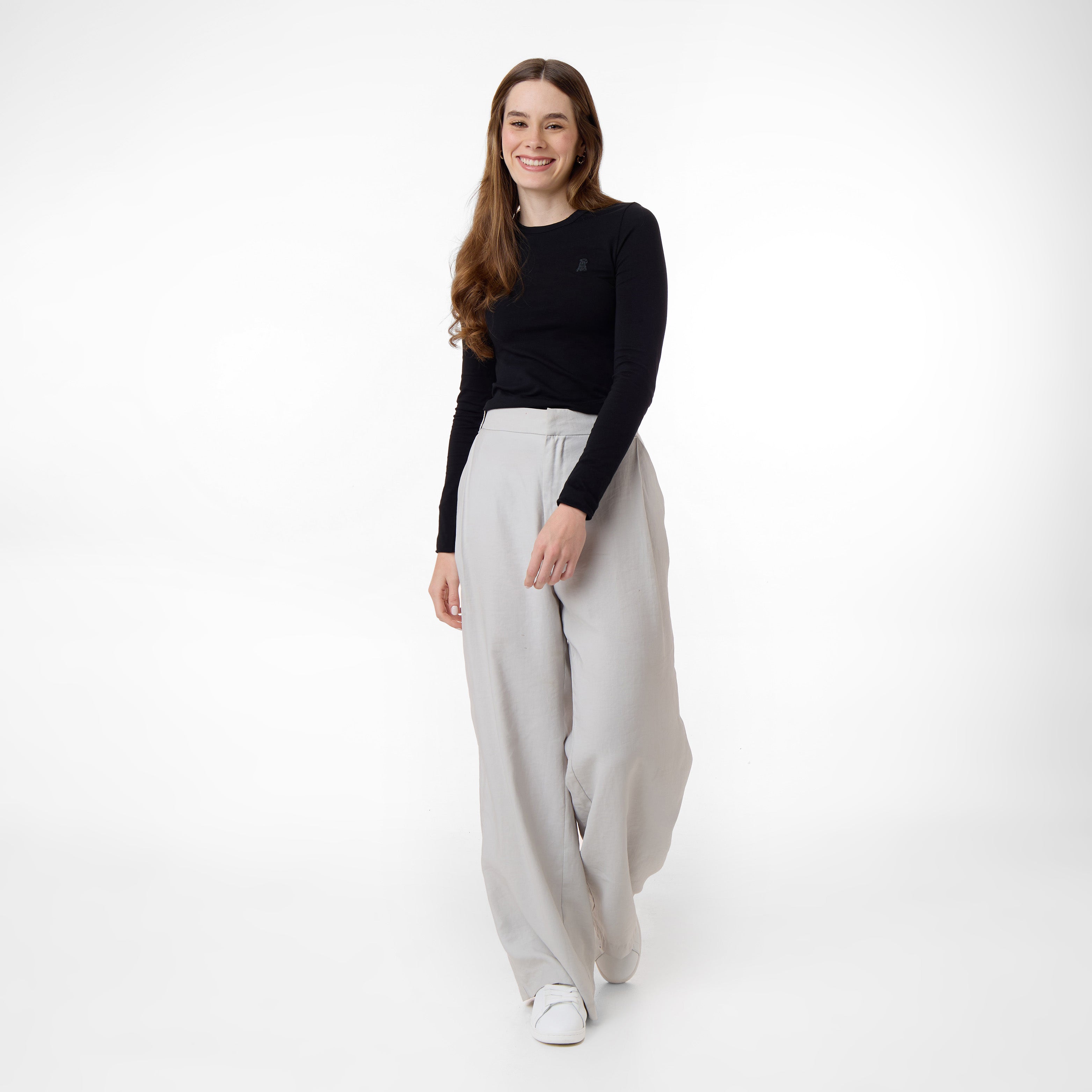 A female model wearing a black long-sleeve top paired with wide-legged light gray trousers. She is walking forward with a smile, exuding a relaxed and confident vibe. The outfit is simple and elegant, set against a white background.