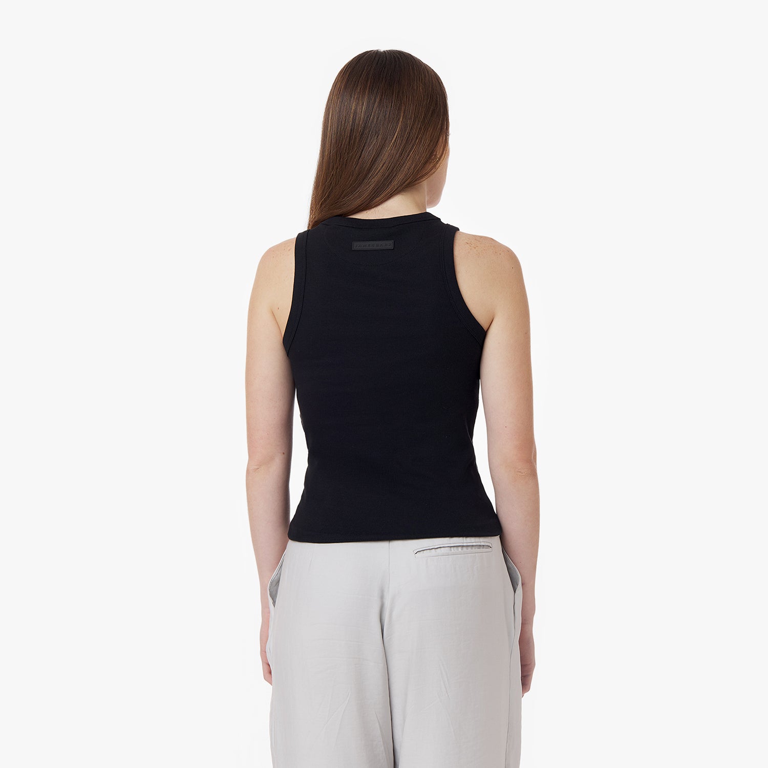 Back view of a female model wearing a black sleeveless ribbed tank top with a fitted silhouette. Features a black rectangular 'JAMES BARK' label near the neckline. Paired with light-colored wide-leg pants. Minimalist white background