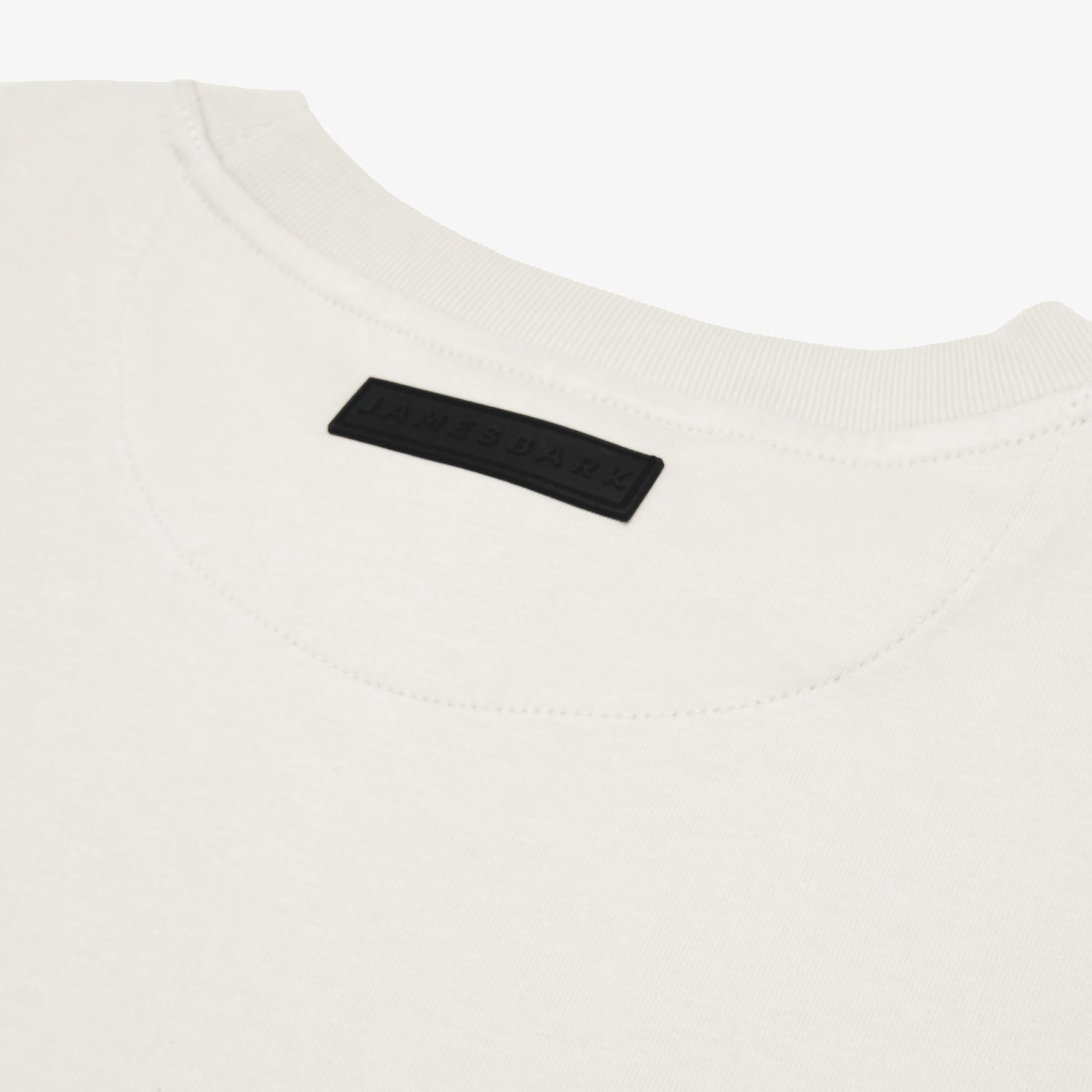 Close-up of the back of a white t-shirt, highlighting a black rectangular 'JAMES BARK' label near the neckline. The fabric appears soft with a ribbed collar and a minimalist aesthetic. White background