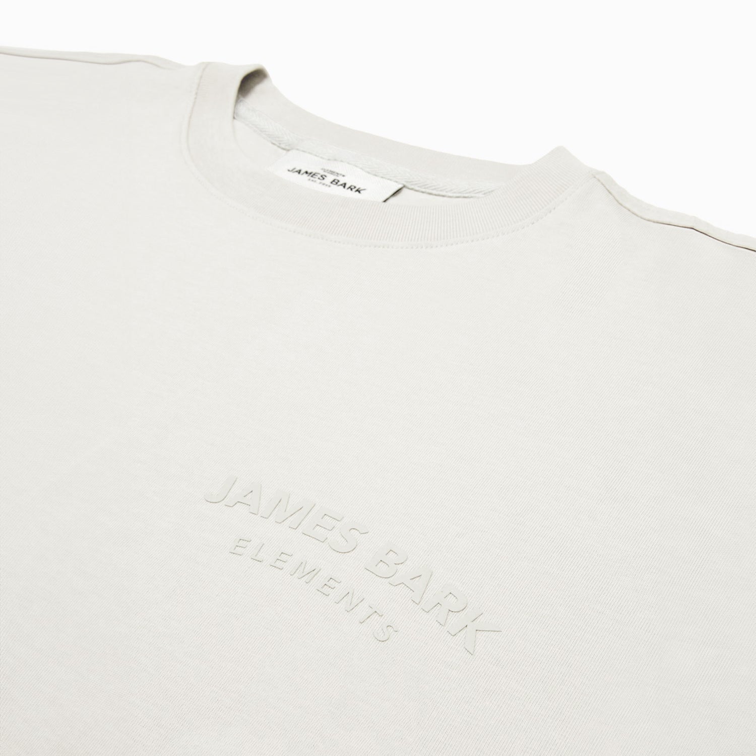 Close-up of a white t-shirt featuring an embossed 'JAMES BARK ELEMENTS' logo on the chest. The fabric appears soft, with a ribbed crew neck collar and a minimalist design. White background