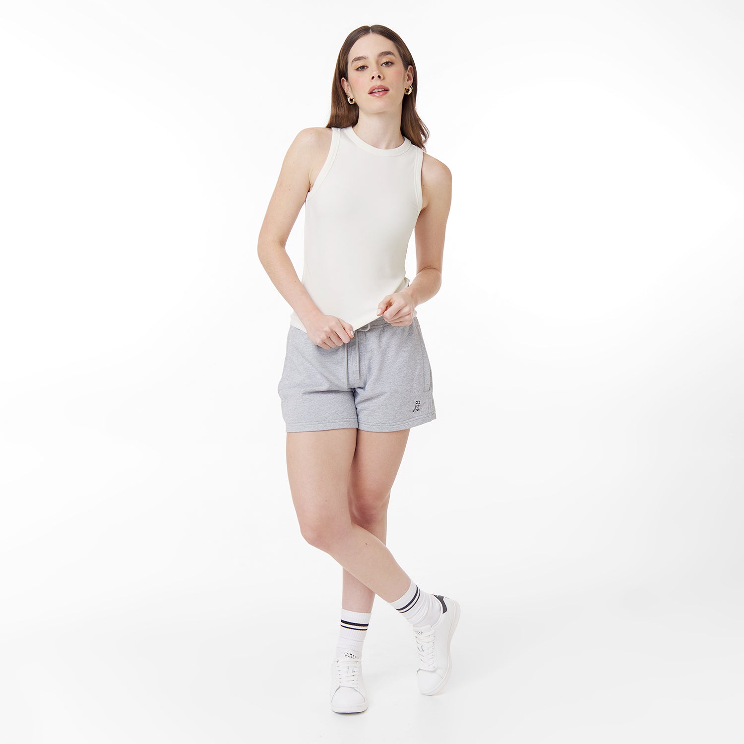 A female model with long brown hair poses in a white fitted ribbed tank top and light gray drawstring shorts with a small embroidered logo. She pairs the outfit with white sneakers and striped socks. The background is plain white.