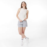 A female model with long brown hair poses in a white fitted ribbed tank top and light gray drawstring shorts with a small embroidered logo. She pairs the outfit with white sneakers and striped socks. The background is plain white.