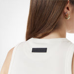 Close-up of a woman's upper back, highlighting a white ribbed tank top with a black rectangular "JAMES BARK" logo patch sewn below the neckline. Her long brown hair is partially covering the top, and she wears multiple gold and gemstone earrings. The background is plain white.