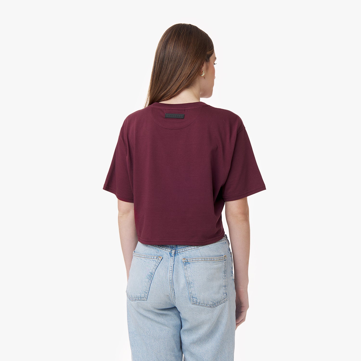 A young woman is seen from the back wearing a burgundy cropped T-shirt with a small black label near the neckline and high-waisted light blue jeans. Her long, straight brown hair falls over her shoulders, and she stands against a plain white background