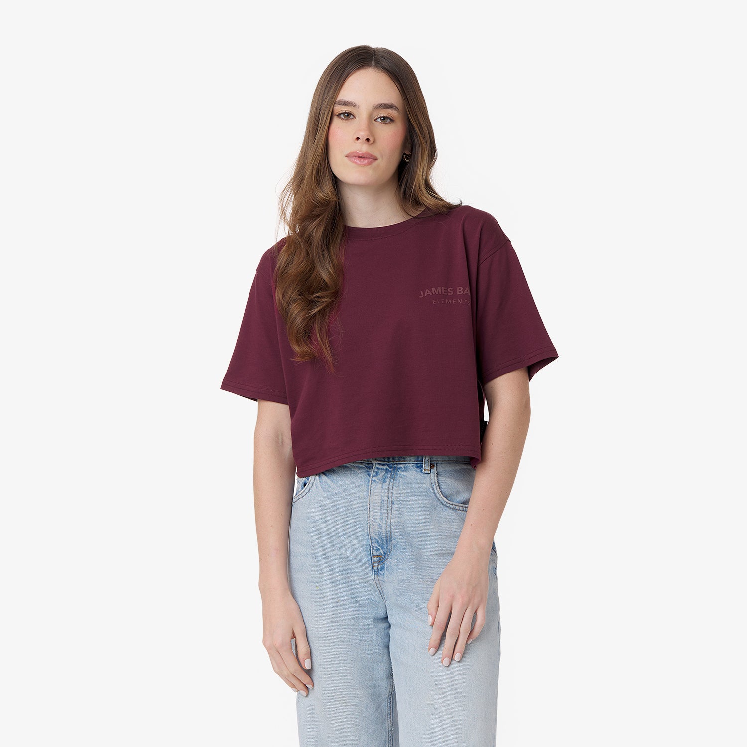 A young woman with long wavy brown hair wears a burgundy cropped T-shirt with small text on the upper left side and high-waisted light blue jeans. She stands against a plain white background with a neutral expression