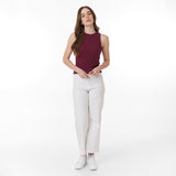 Full-body shot of a woman with long, wavy brown hair wearing a burgundy sleeveless tank top and high-waisted white jeans with a frayed hem. She pairs the outfit with white sneakers. She poses with a relaxed stance, slightly tilting her head, with her hands near the button of her jeans. The background is a clean white backdrop.