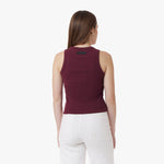 A woman with long brown hair, wearing gold hoop earrings, is shown from the back wearing a burgundy sleeveless ribbed tank top with a black "JAMES BARK" logo patch on the upper back. She pairs it with high-waisted white pants that feature two back pockets. The background is plain white