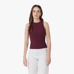 A woman with long brown hair and gold hoop earrings is wearing a sleeveless, ribbed burgundy tank top with a high neckline, paired with high-waisted white pants. She stands against a plain white background with a neutral expression.