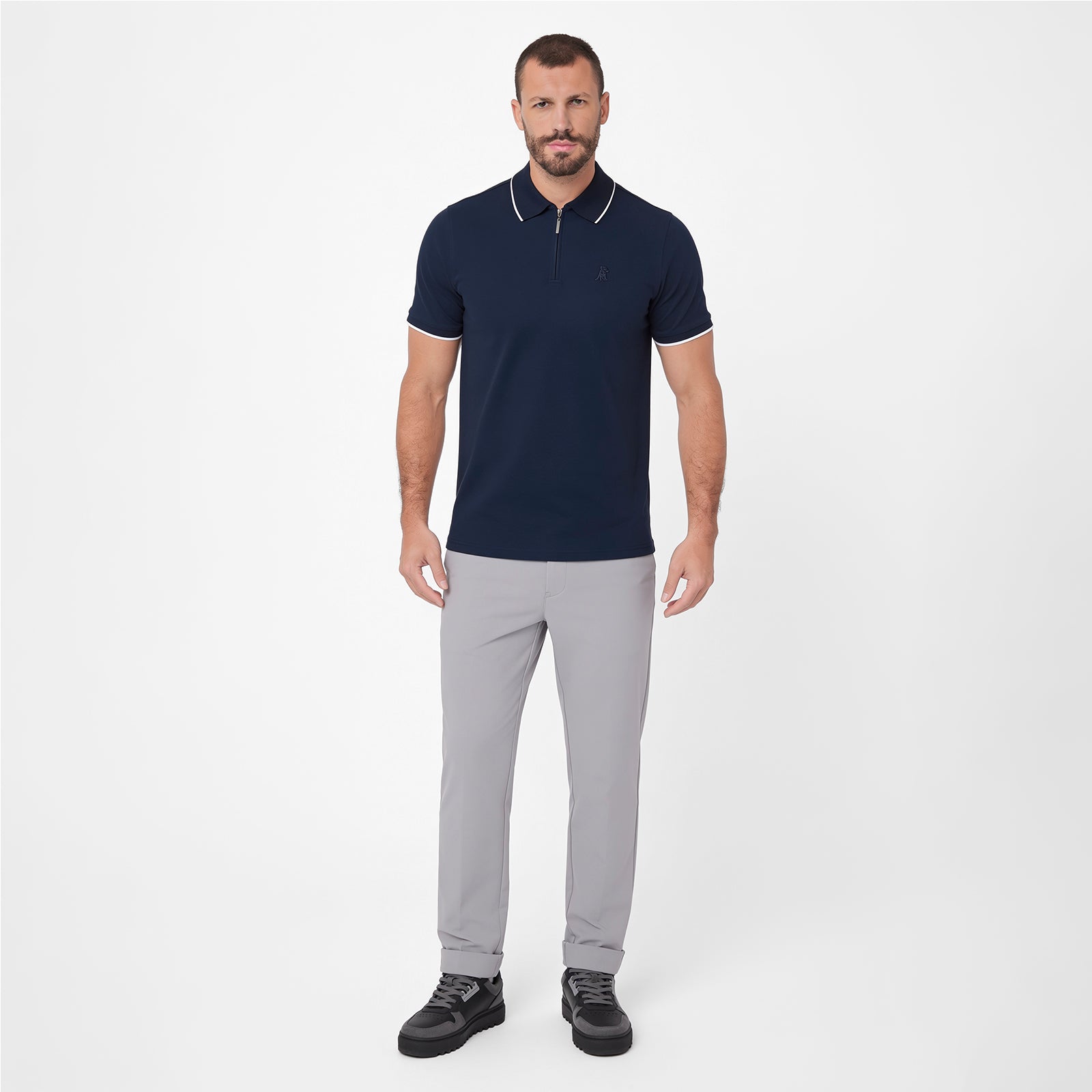 A man modeling a short-sleeve polo shirt in navy blue with white trim on the collar and sleeve cuffs, paired with light gray trousers and black sneakers