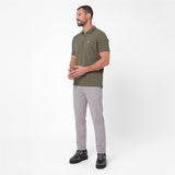 A man wearing an olive green short-sleeve polo shirt, light gray trousers, and black sneakers, standing against a plain white studio background.