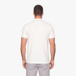 A man viewed from the back wearing a short-sleeve polo shirt in off-white and light gray trousers