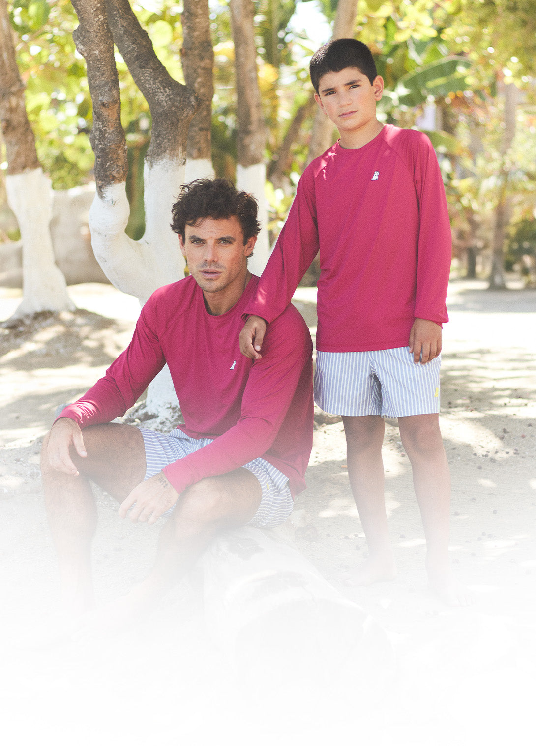 Two people wearing pink long-sleeve shirts and white shorts sitting on a sandy beach with trees in the background.