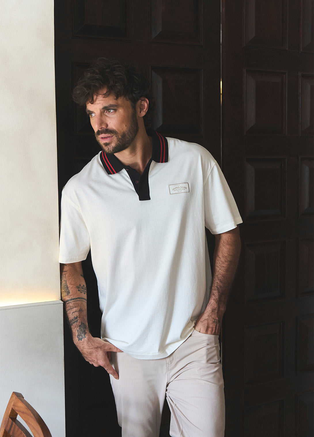 Man wearing a white James Bark short-sleeve polo shirt with a contrasting black collar featuring red stripes and a beige chest logo patch