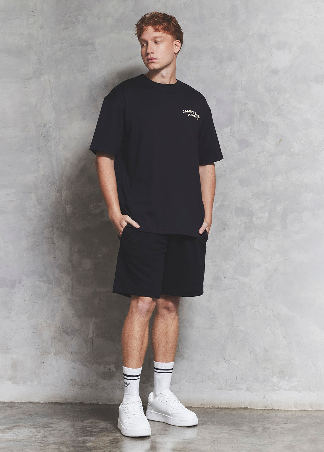 Man wearing a black t-shirt and shorts with white sneakers against a gray concrete wall.