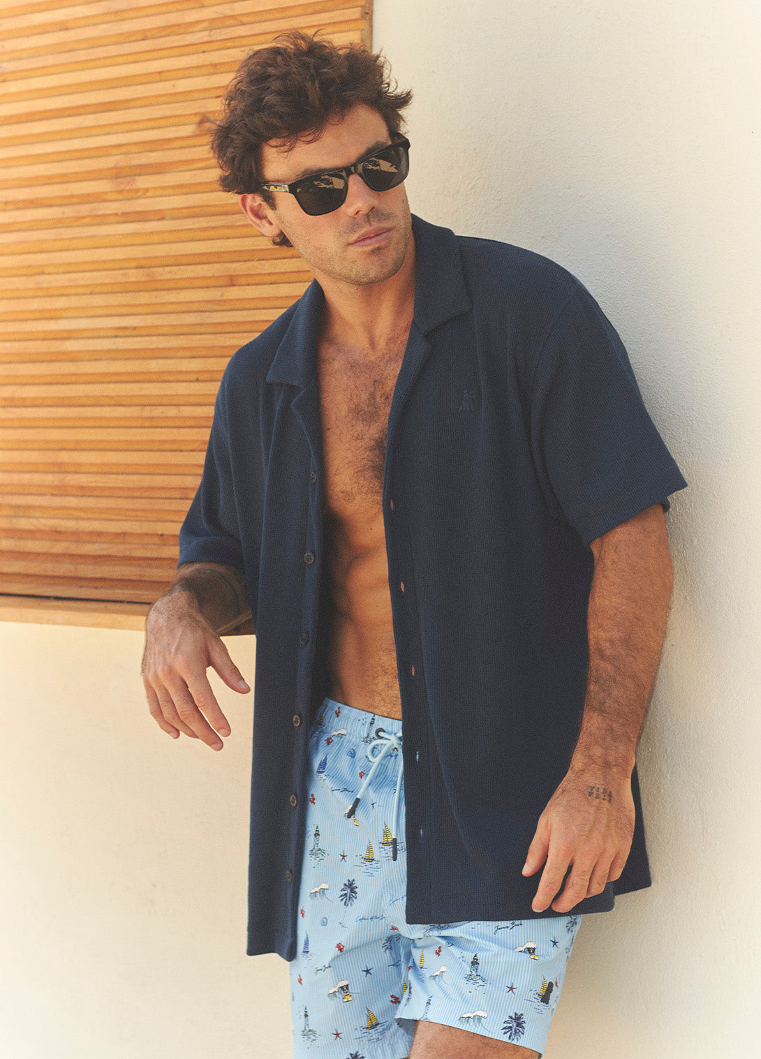 Man wearing sunglasses and a navy shirt with light blue patterned shorts against a neutral wall.
