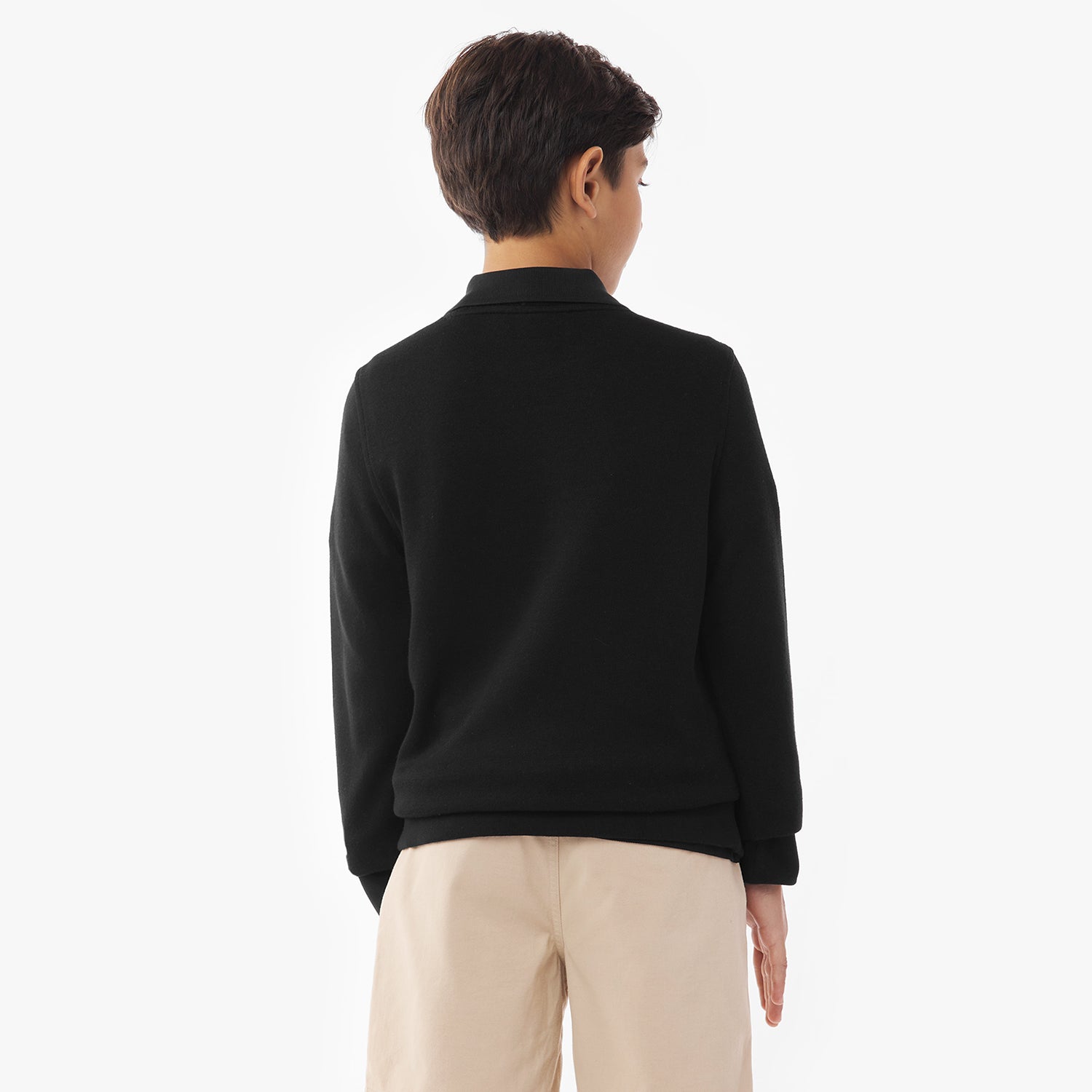 A young boy shown from the back, wearing a black sweater over a collared shirt and beige shorts. His hands are relaxed by his sides, and the background is plain white.