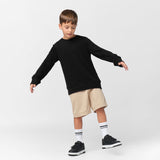 A young boy with short brown hair balances playfully with his arms outstretched, wearing a black oversized long-sleeve sweatshirt with wide cuffs, beige shorts, white crew socks with black stripes, and black sneakers with velcro straps. The sweatshirt features a subtle embroidered dog logo on the left chest. He has a focused expression and is looking downward. The background is plain white.