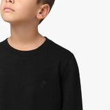 A close-up of a young boy wearing a black long-sleeve sweatshirt with a ribbed crew neckline. The sweatshirt features a small embroidered black-on-black dog logo on the left chest. The fabric has a subtle ribbed texture. The boy’s face is partially visible, showing his chin and lips as he looks slightly upward. The background is plain white.