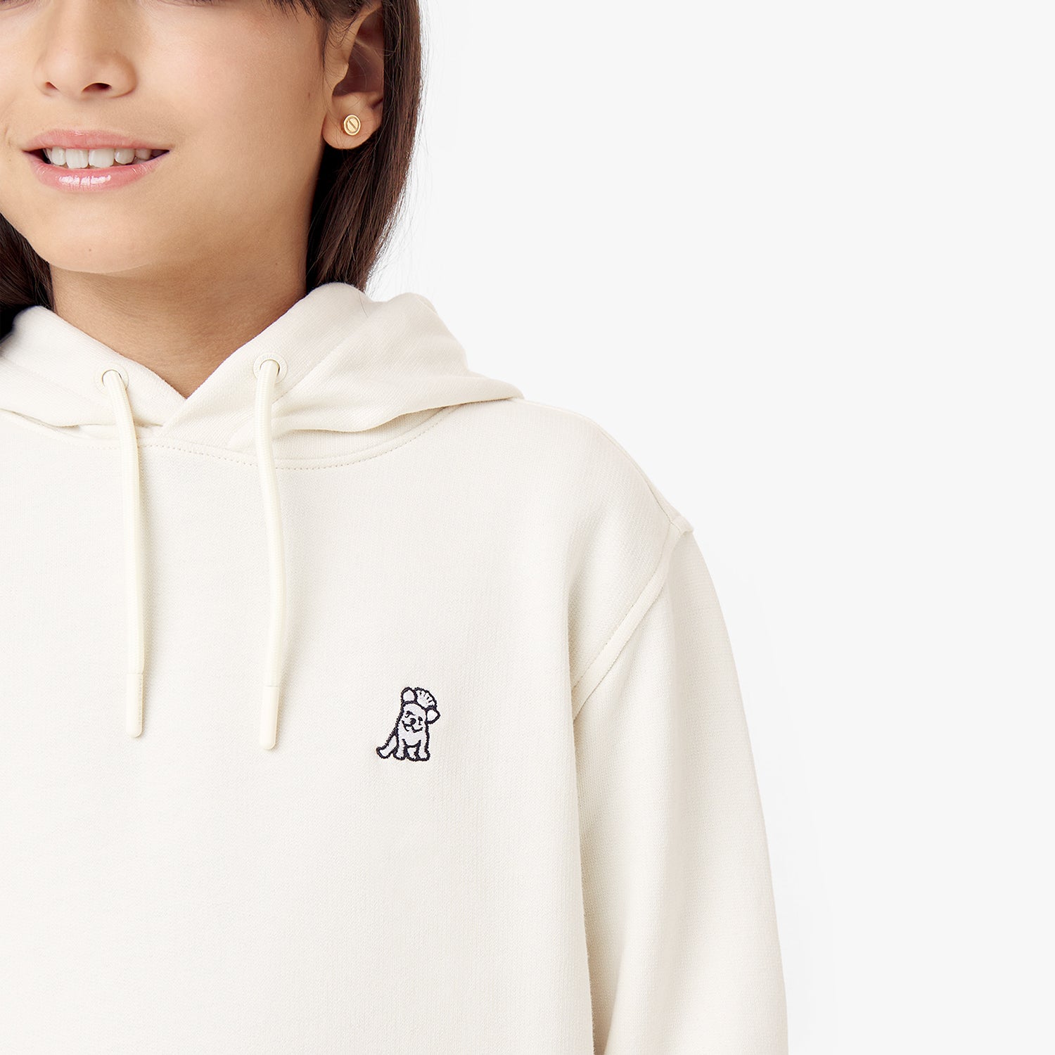 Close-up side view of the cream-colored hoodie worn by a child, focusing on the chest area with the embroidered dog logo and the texture of the fabric.