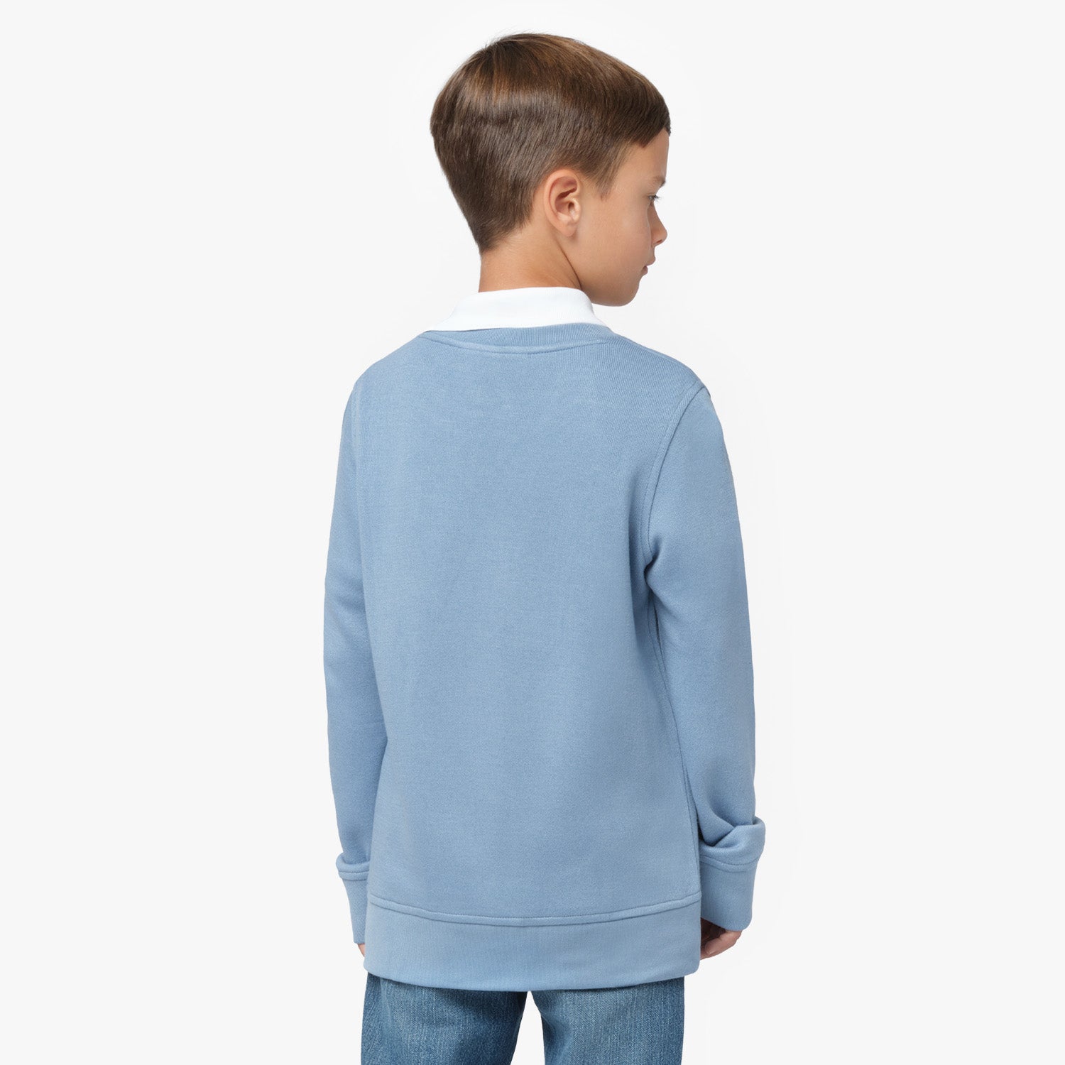 A young boy with short brown hair is shown from the back, wearing a light blue oversized sweatshirt with a white collared shirt underneath. The sweatshirt has a relaxed fit with wide cuffs. He is also wearing blue jeans. His head is slightly turned to the side, showing a partial profile. The background is plain white.