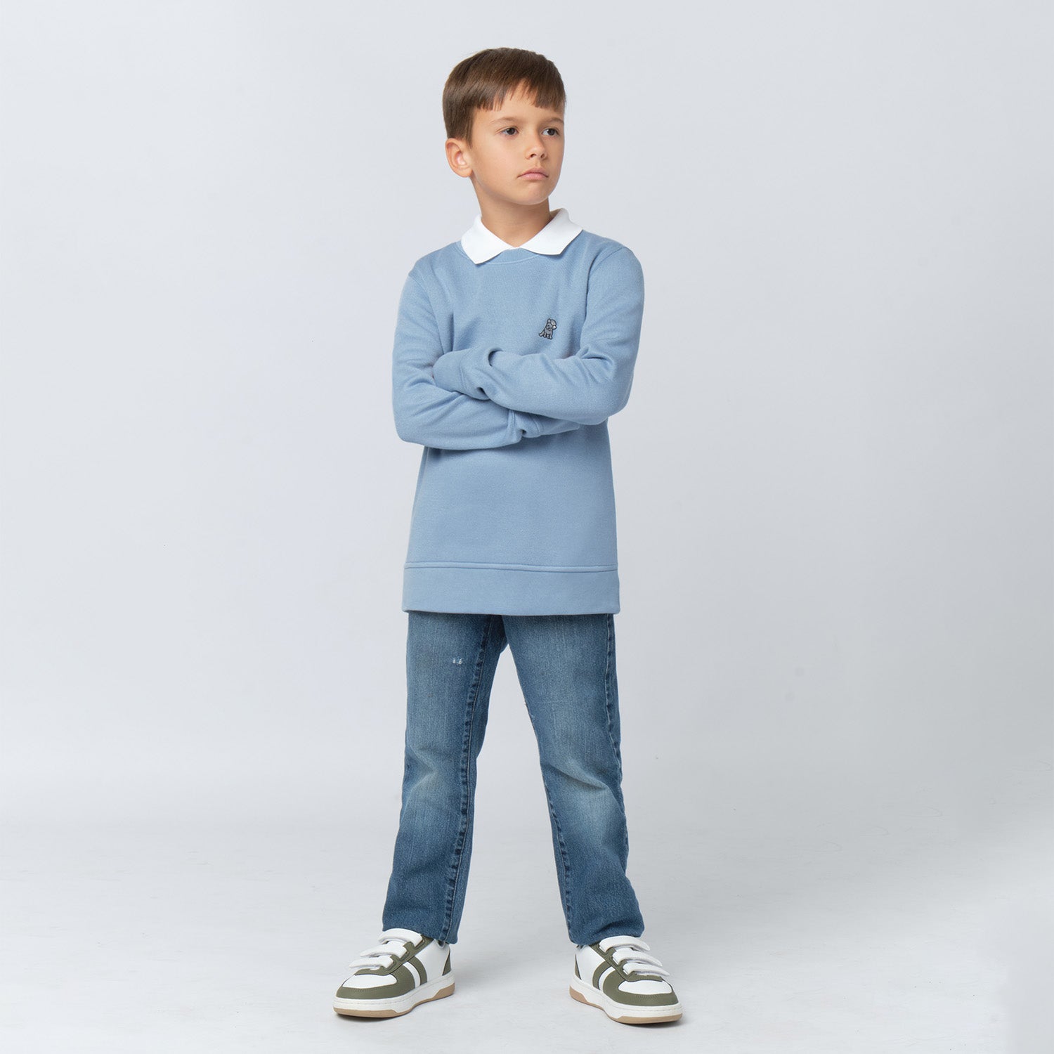 A young boy with short brown hair stands with his arms crossed, wearing a light blue oversized sweatshirt with a white collared shirt underneath. The sweatshirt features a small embroidered dog logo on the left chest. He pairs it with blue jeans and white sneakers with green and brown accents. He has a confident expression and looks to the side against a light gray background.