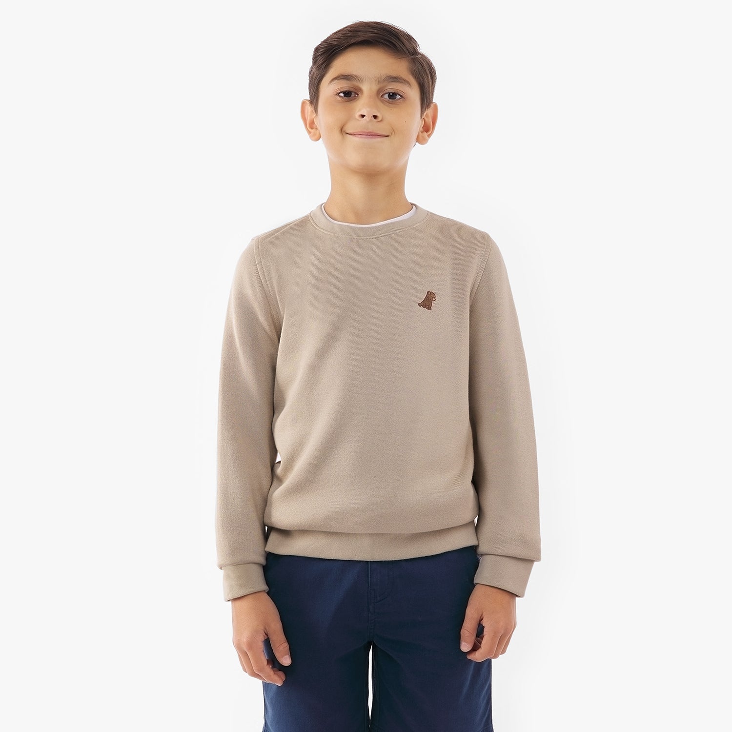 A young boy facing forward, wearing a beige sweatshirt with a small embroidered logo on the left chest and navy blue shorts. He has a slight smile and stands in a relaxed posture.