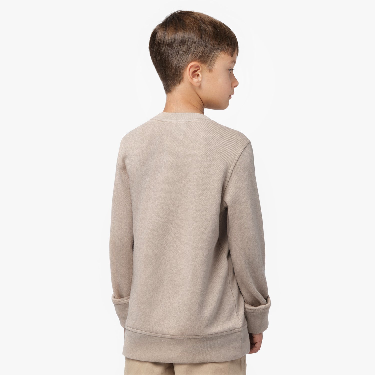 A young boy with short brown hair is shown from the back, wearing a beige oversized long-sleeve sweatshirt. The sweatshirt has a relaxed fit with wide cuffs and a slightly textured fabric. His head is slightly turned to the side, revealing a partial profile. The background is plain white.